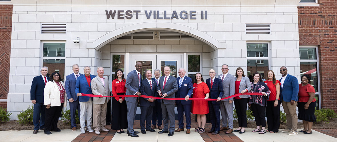 A group of people stands in front of a brick building labeled "WEST VILLAGE II," participating in a ribbon-cutting ceremony.